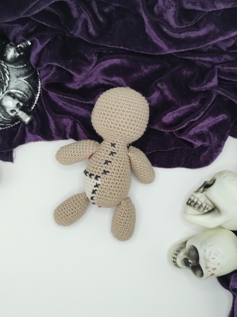 Puede incluir: Una mu&ntilde;eca vud&uacute; de crochet con costuras negras y un parche de tela marr&oacute;n en la espalda. La mu&ntilde;eca est&aacute; tumbada sobre una superficie blanca con dos cr&aacute;neos de pl&aacute;stico blanco.