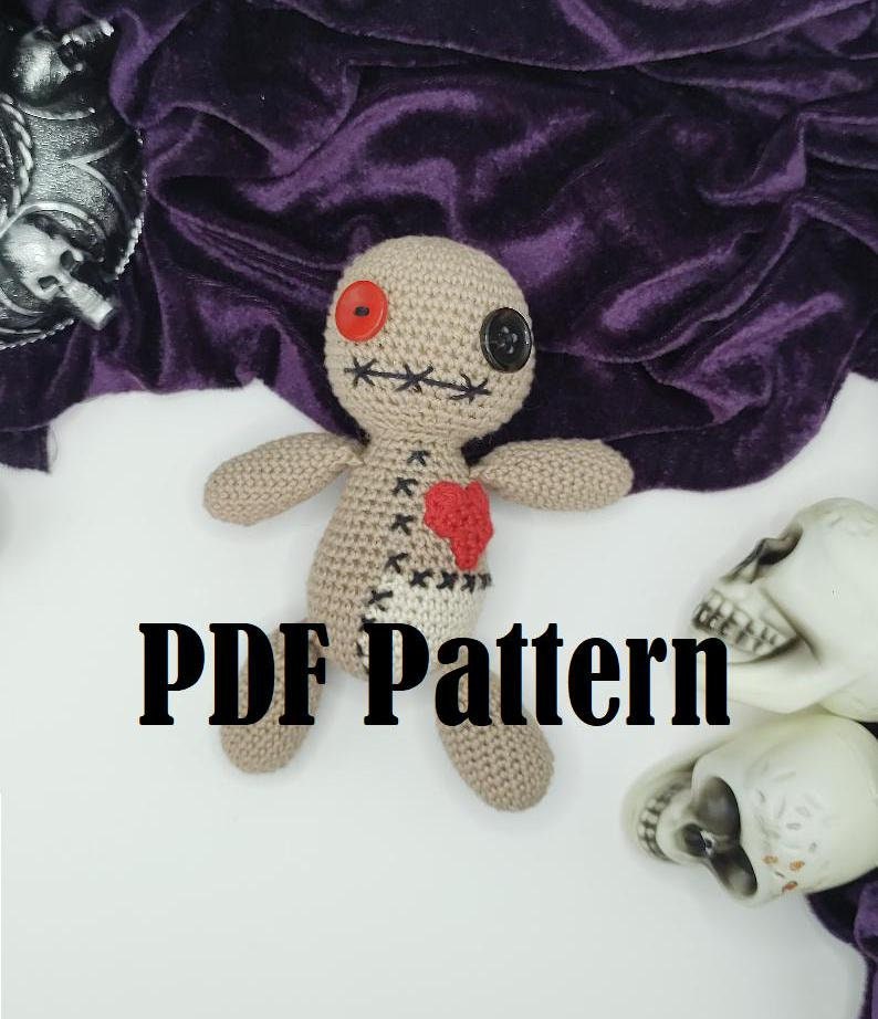 Puede incluir: Patr&oacute;n de crochet para un mu&ntilde;eco vud&uacute; con un ojo de bot&oacute;n rojo, un ojo de bot&oacute;n negro y un coraz&oacute;n rojo en el pecho. El mu&ntilde;eco est&aacute; tumbado sobre una superficie blanca con dos cr&aacute;neos blancos en el fondo. El texto "PDF Pattern" est&aacute; impreso en negro sobre la superficie blanca.