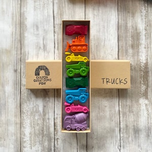 May include: A set of 8 crayon truck and car shaped crayons in a cardboard box. The crayons are arranged in a rainbow order from red to purple. The box is labeled "Crayon Creations PDX" and "Trucks".