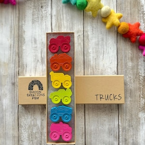 May include: A set of six colourful monster truck crayons in a cardboard box. The box is labelled "Crayon Creations PDX" and "Trucks".