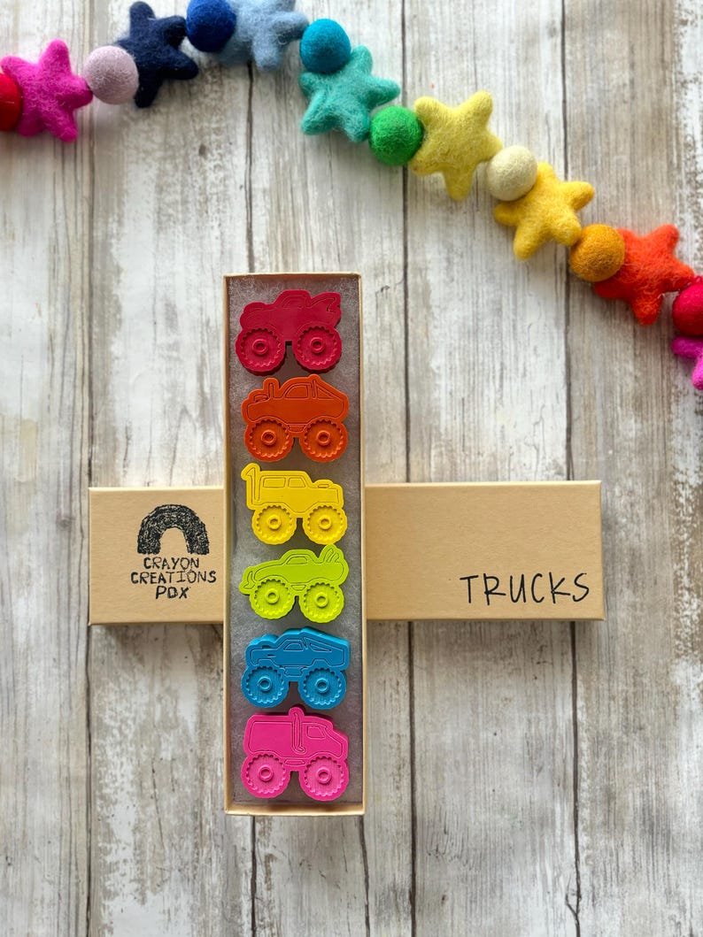 May include: A set of six colourful monster truck crayons in a cardboard box. The box is labelled "Crayon Creations PDX" and "Trucks".