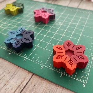 Puede incluir: Cuatro crayones con forma de copo de nieve en diferentes colores: verde, rojo, azul y naranja. Los crayones est&aacute;n sobre una estera de corte verde con marcas blancas.