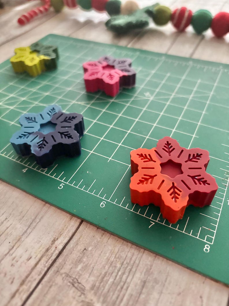 Puede incluir: Cuatro crayones con forma de copo de nieve en diferentes colores: verde, rojo, azul y naranja. Los crayones est&aacute;n sobre una estera de corte verde con marcas blancas.