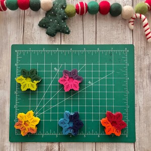 Puede incluir: Cinco adornos de madera con forma de copo de nieve de colores sobre una estera de corte verde. Los copos de nieve est&aacute;n pintados en tonos de rojo, naranja, amarillo, azul y verde.