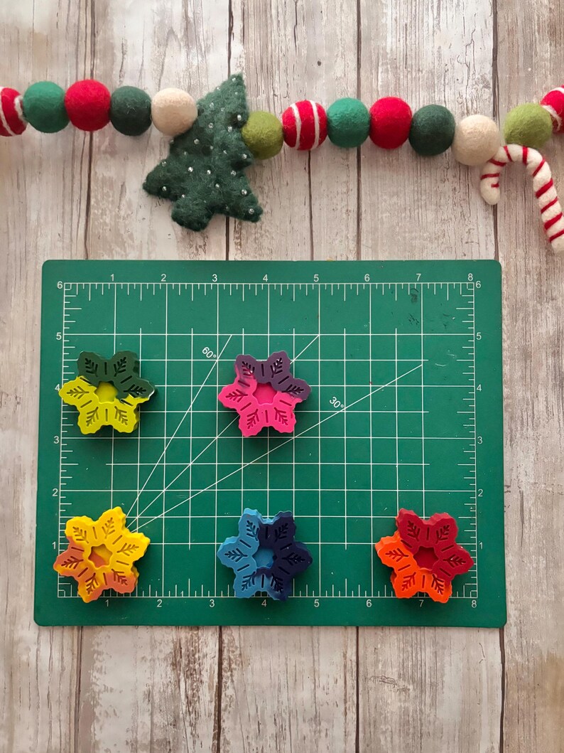 Puede incluir: Cinco adornos de madera con forma de copo de nieve de colores sobre una estera de corte verde. Los copos de nieve est&aacute;n pintados en tonos de rojo, naranja, amarillo, azul y verde.