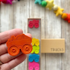 May include: Orange monster truck shaped rubber stamp with a textured surface. The stamp is held in a hand. The stamp is part of a set of four stamps in a wooden box labelled "TRUCKS".