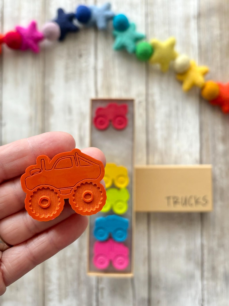 May include: Orange monster truck shaped rubber stamp with a textured surface. The stamp is held in a hand. The stamp is part of a set of four stamps in a wooden box labelled "TRUCKS".