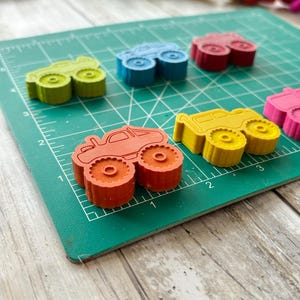 May include: A set of colourful monster truck shaped crayons on a green cutting mat. The crayons are red, yellow, blue, green, and orange.