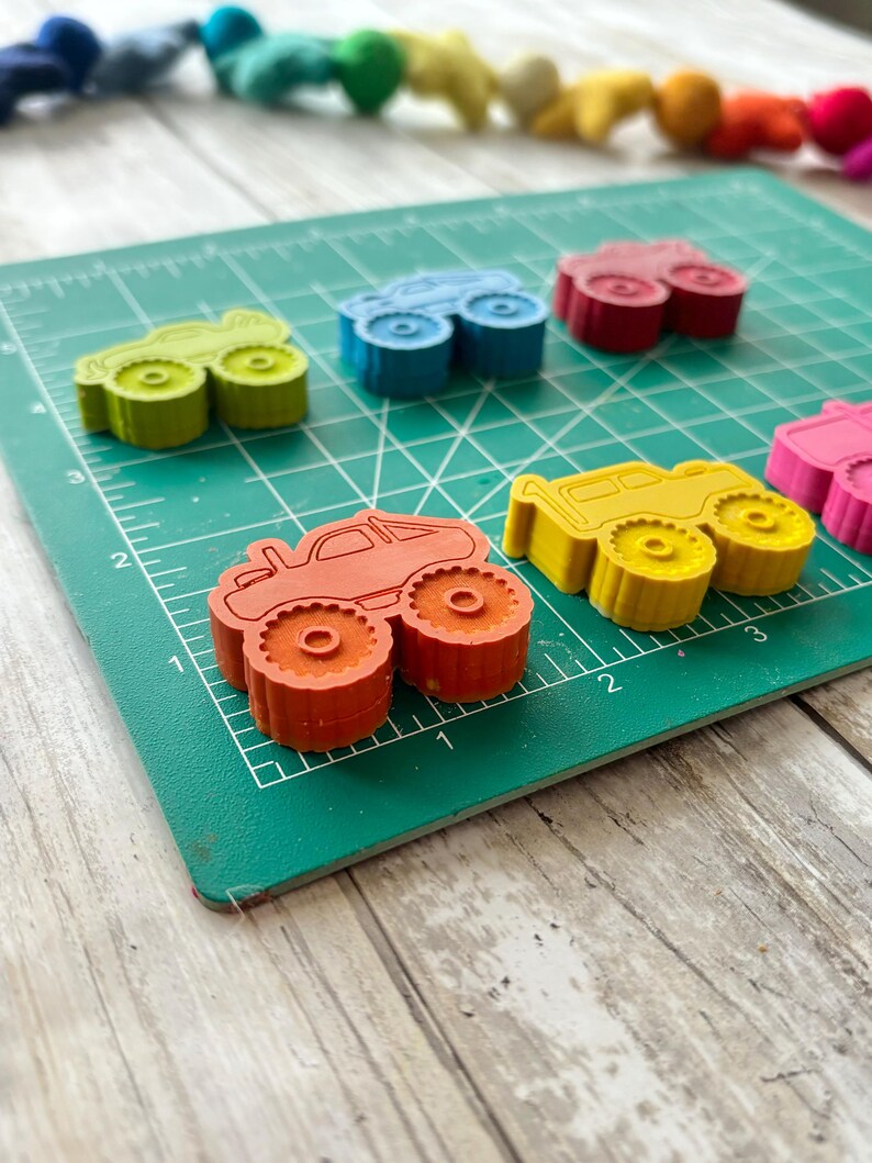 May include: A set of colourful monster truck shaped crayons on a green cutting mat. The crayons are red, yellow, blue, green, and orange.