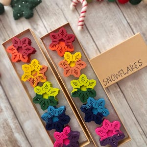 Puede incluir: Dos cajas de crayones con forma de copo de nieve de color arco&iacute;ris. Las cajas son de cart&oacute;n y de color marr&oacute;n. El texto "SNOWFLAKES" est&aacute; impreso en la parte superior de una de las cajas.