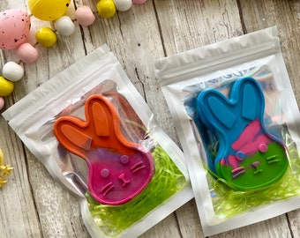 Könnte beinhalten: Zwei bunte Hasen-förmige Wachsmalstifte in einzelnen durchsichtigen Plastiktüten mit grünem zerkleinertem Papier. Die Wachsmalstifte sind ein rosa-orangener Farbverlauf und ein blau-rosa-grüner Farbverlauf.