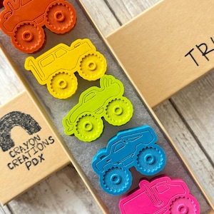 May include: A set of six monster truck crayons in a rainbow of colours, including red, orange, yellow, green, blue, and pink. The crayons are in a box with the word "TRUCK" written on it.