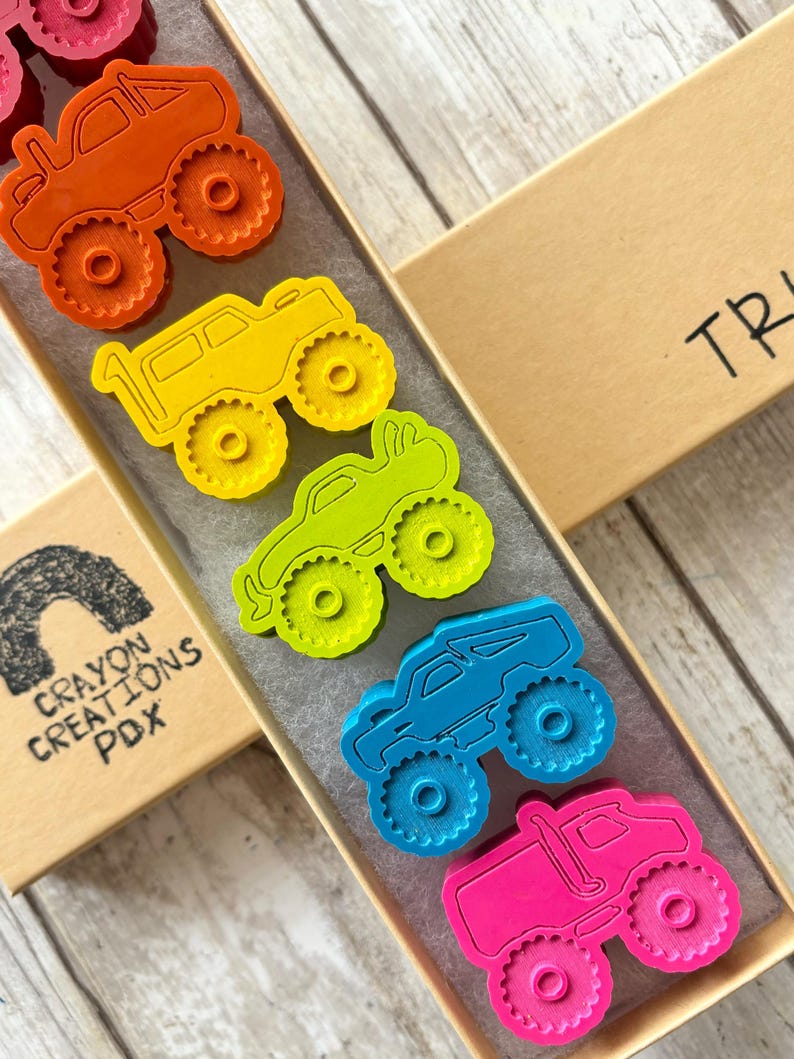 May include: A set of six monster truck crayons in a rainbow of colours, including red, orange, yellow, green, blue, and pink. The crayons are in a box with the word "TRUCK" written on it.