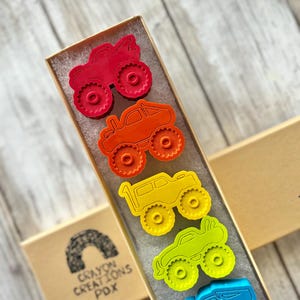 May include: A set of seven monster truck cookie cutters in a rainbow of colours, including red, orange, yellow, green, blue, and purple. The cookie cutters are arranged in a row in a box with the text "Crayon Creations PDX" on the side.