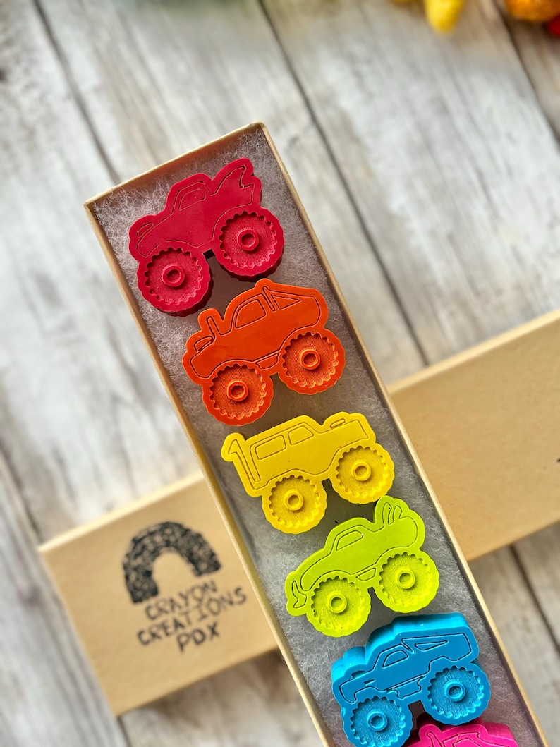May include: A set of seven monster truck cookie cutters in a rainbow of colours, including red, orange, yellow, green, blue, and purple. The cookie cutters are arranged in a row in a box with the text "Crayon Creations PDX" on the side.
