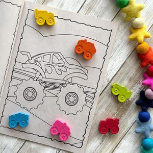May include: A colouring book page featuring a monster truck illustration.  The page is surrounded by small toy monster trucks in various colours, including yellow, orange, red, pink, blue and green.