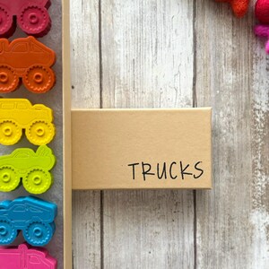 May include: A box of colourful monster truck shaped crayons. The box is labelled "TRUCKS".