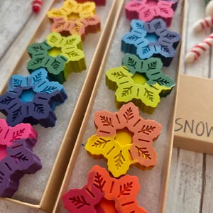 Puede incluir: Dos cajas llenas de crayones con forma de copo de nieve de colores arco&iacute;ris. Las cajas son de cart&oacute;n y tienen un interior blanco. Los crayones est&aacute;n dispuestos en un patr&oacute;n de arco&iacute;ris, con el cray&oacute;n rojo en la parte superior y el cray&oacute;n morado en la parte inferior. Las cajas est&aacute;n etiquetadas con "SNOW".