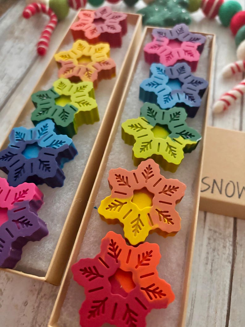 Puede incluir: Dos cajas llenas de crayones con forma de copo de nieve de colores arco&iacute;ris. Las cajas son de cart&oacute;n y tienen un interior blanco. Los crayones est&aacute;n dispuestos en un patr&oacute;n de arco&iacute;ris, con el cray&oacute;n rojo en la parte superior y el cray&oacute;n morado en la parte inferior. Las cajas est&aacute;n etiquetadas con "SNOW".