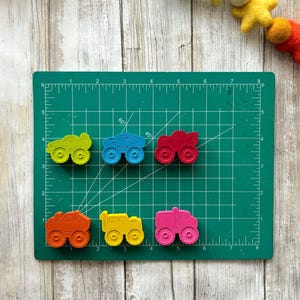 May include: Six colourful toy monster trucks on a green cutting mat. The trucks are yellow, blue, red, orange, yellow, and pink. The trucks have black wheels and are made of plastic.