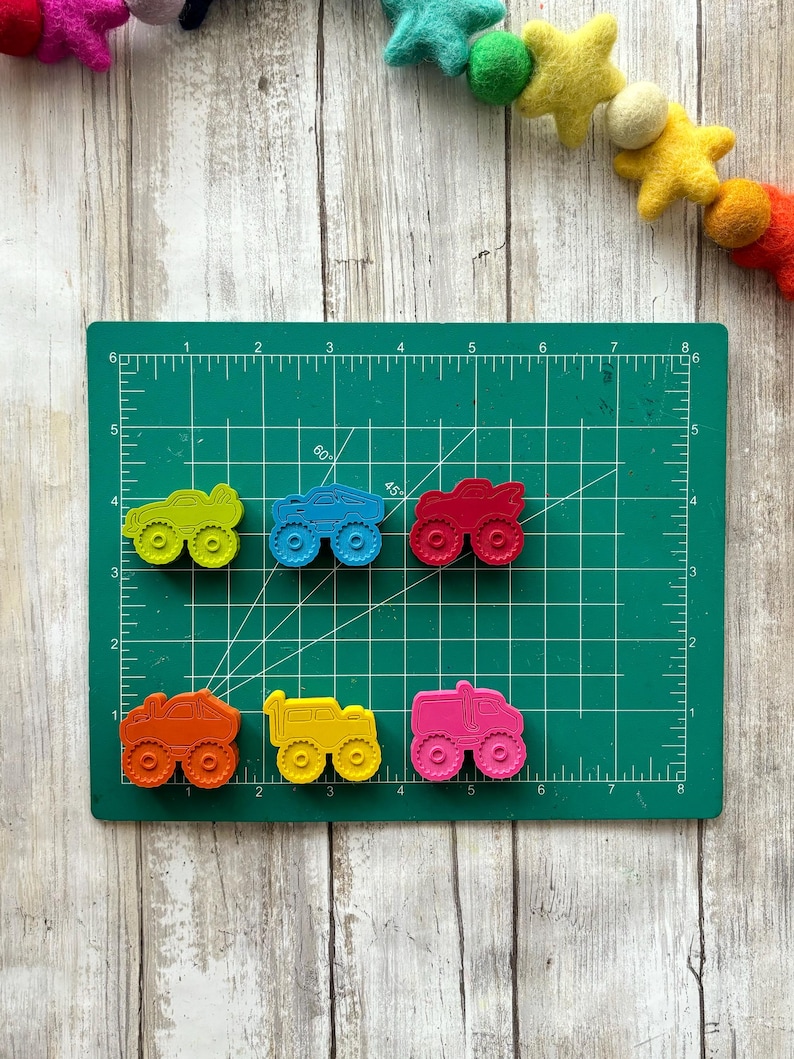 May include: Six colourful toy monster trucks on a green cutting mat. The trucks are yellow, blue, red, orange, yellow, and pink. The trucks have black wheels and are made of plastic.
