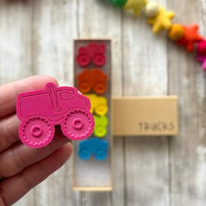 May include: A pink monster truck toy with large wheels. The truck is part of a set of colourful toy trucks in a wooden box labelled "TRUCKS".