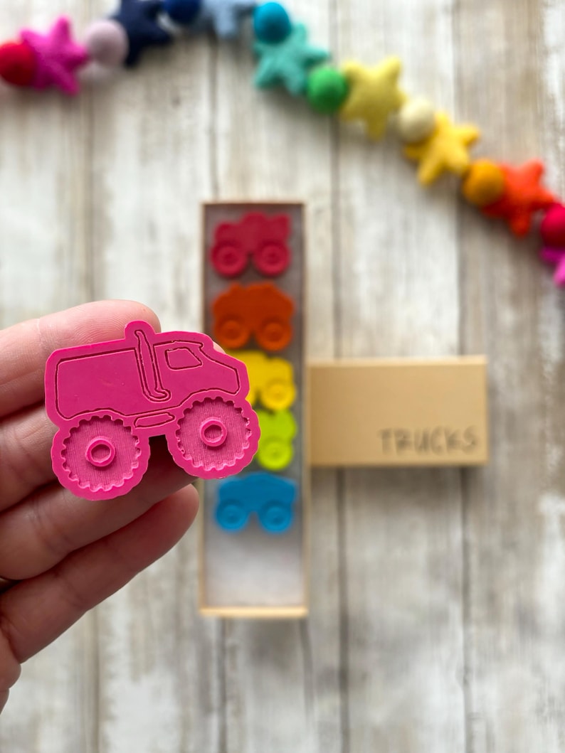 May include: A pink monster truck toy with large wheels. The truck is part of a set of colourful toy trucks in a wooden box labelled "TRUCKS".