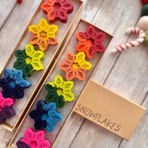 Puede incluir: Un conjunto de diez crayones de colores en forma de copo de nieve en una caja marr&oacute;n con la palabra "SNOWFLAKES" escrita en ella.