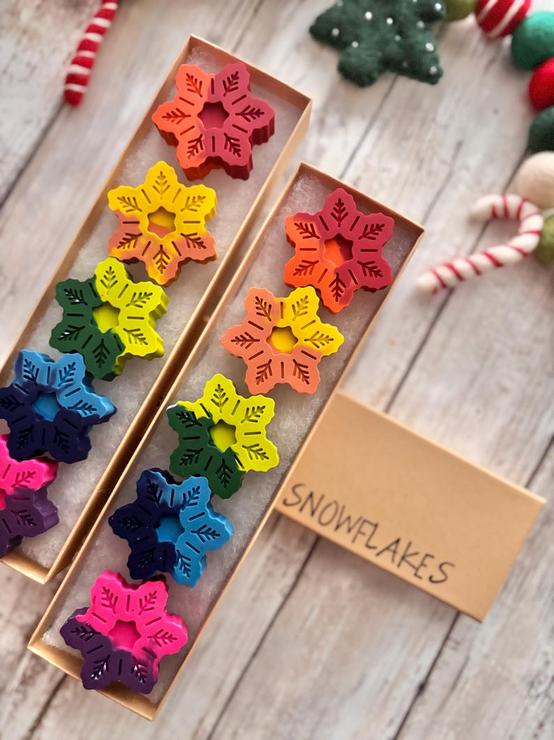 Puede incluir: Un conjunto de diez crayones de colores en forma de copo de nieve en una caja marr&oacute;n con la palabra "SNOWFLAKES" escrita en ella.