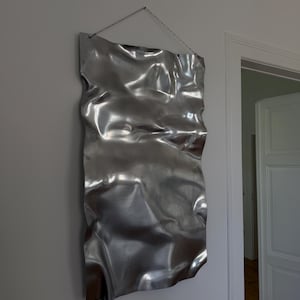 Könnte beinhalten: Ein großer, rechteckiger, metallischer Wandbehang mit zerknitterter, strukturierter Oberfläche. Das silberfarbene Kunstwerk ist an einer Kette aufgehängt und an einer weißen Wand befestigt. Die reflektierende Oberfläche des Kunstwerks erzeugt Glanzlichter und Schatten.