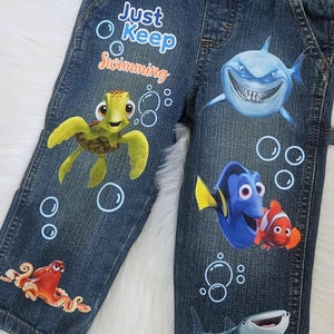 May include: Blue denim overalls featuring Finding Nemo characters including a sea turtle, shark, and clownfish. The text reads Finding Brayzen and Just Keep Swimming, accompanied by white bubble graphics printed across the front of the garment.