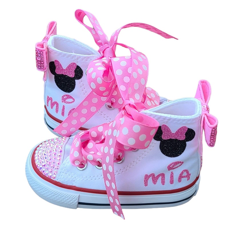 Puede incluir: Zapatillas blancas de ca&ntilde;a alta con cordones de cinta rosa con lunares blancos y un dise&ntilde;o de Minnie Mouse negro y rosa. Las zapatillas est&aacute;n decoradas con pedrer&iacute;a y el nombre "MIA" est&aacute; escrito en el lateral.