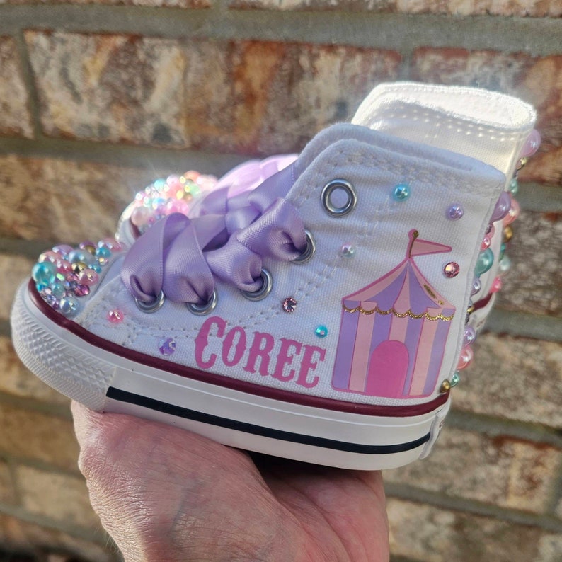 May include: White high-top sneakers with purple ribbon laces and a pink and purple circus tent design. The sneakers are decorated with rhinestones and the name "Coree" is written on the side.