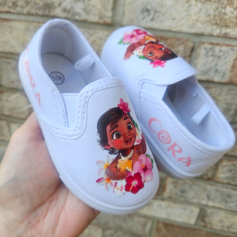 May include: White canvas slip-on shoes with a pink floral design and a cartoon image of Moana from Disney's Moana. The shoes are personalized with the names "Cora" and "Ada" in pink.