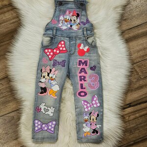 Puede incluir: Peto vaquero azul claro con dise&ntilde;os de Minnie Mouse y Daisy Duck, lazos y el nombre "MARLO" y el n&uacute;mero "3" en purpurina rosa. El peto tiene tirantes ajustables y bolsillos.