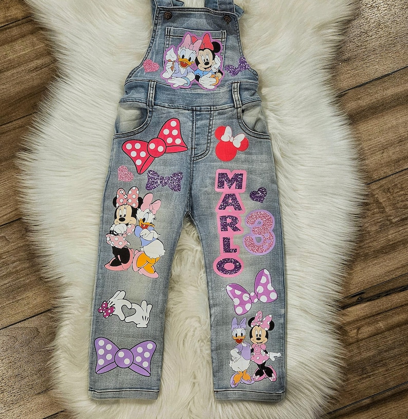 Puede incluir: Peto vaquero azul claro con dise&ntilde;os de Minnie Mouse y Daisy Duck, lazos y el nombre "MARLO" y el n&uacute;mero "3" en purpurina rosa. El peto tiene tirantes ajustables y bolsillos.