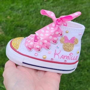 May include: White canvas sneakers with pink polka dot ribbon laces and a gold glitter Minnie Mouse head with the name "Kamille" written below.