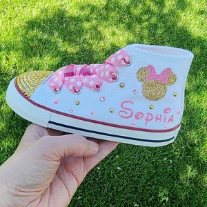May include: White high-top trainers with pink polka dot ribbon laces, gold glitter Minnie Mouse ears, and the name "Sophia" written in pink glitter.