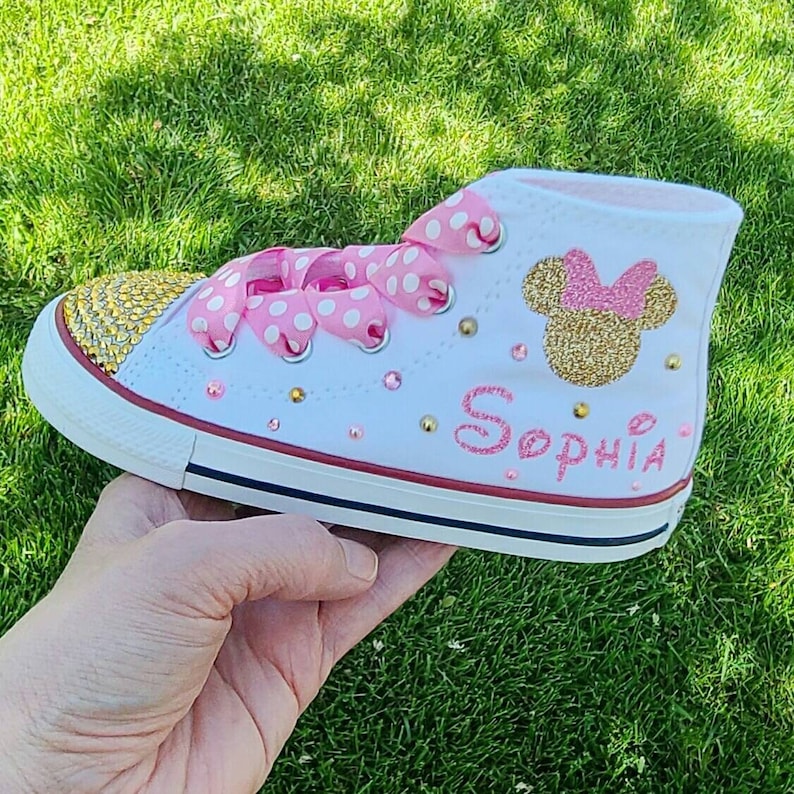 May include: White high-top trainers with pink polka dot ribbon laces, gold glitter Minnie Mouse ears, and the name "Sophia" written in pink glitter.