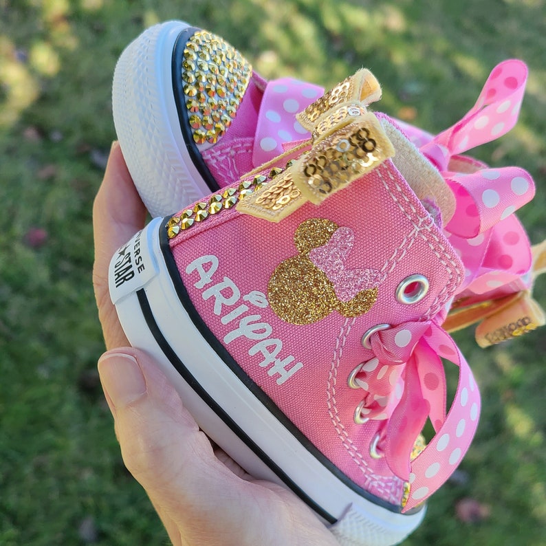 May include: Pink and white high-top Converse sneakers with a gold glitter Minnie Mouse design and the name "Ariyah" written on the side.