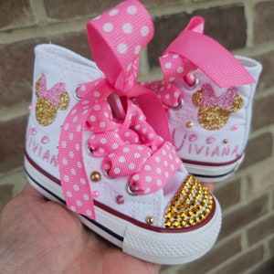 May include: White high-top trainers with pink polka dot ribbon laces and a gold glitter Minnie Mouse design. The shoes are decorated with rhinestones and the name "Vivianna" is written on the side.