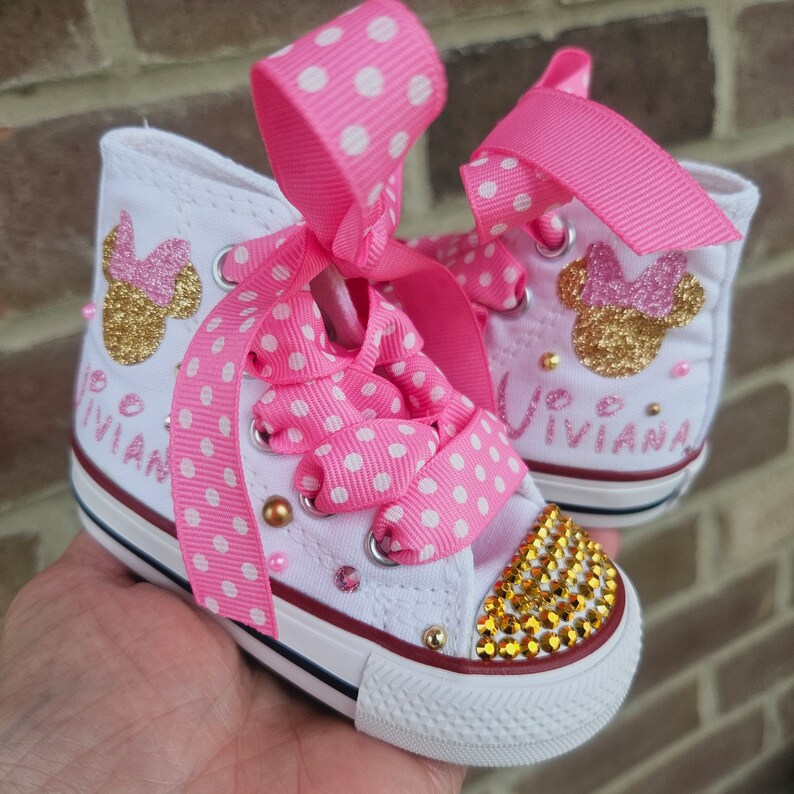 May include: White high-top trainers with pink polka dot ribbon laces and a gold glitter Minnie Mouse design. The shoes are decorated with rhinestones and the name "Vivianna" is written on the side.