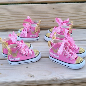 May include: Four pairs of pink high-top sneakers with gold glitter and bows. The sneakers are personalized with the names "Dream", "Arya", "Nevaeh", and a fourth name that is unclear.