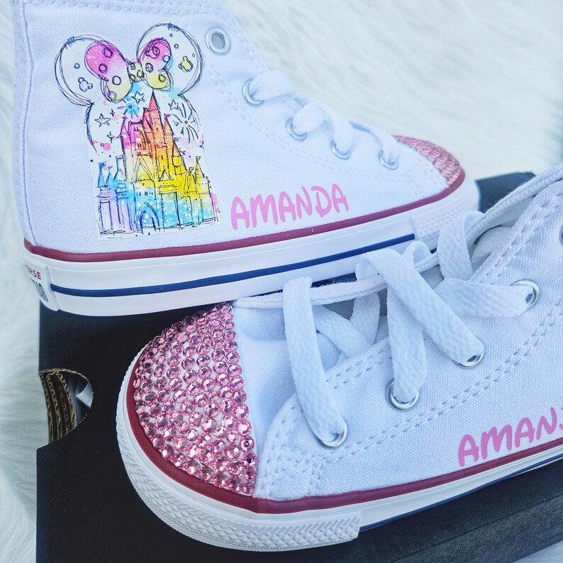 May include: White high-top trainers with pink rhinestone-covered toes. One trainer features a colourful castle design and the name "AMANDA" in pink. The other trainer also has the name "AMANDA" in pink.