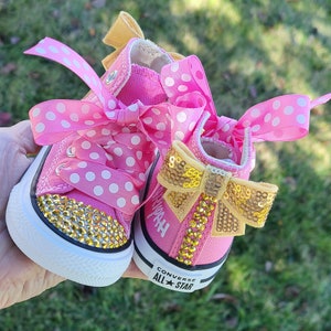 May include: Pink baby Converse sneakers with white polka dots and gold glitter. The shoes have a gold bow with sequins and the words "Converse All Star" on the bottom.