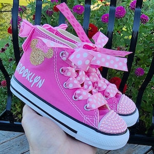 May include: Pink high-top sneakers with white polka dot ribbon laces, a gold glitter Minnie Mouse ear appliqu&eacute;, and the name "Brooklyn" written in white on the side.