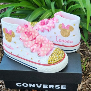 May include: White high-top Converse sneakers with pink and white polka dot ribbon laces. The shoes are decorated with gold glitter Minnie Mouse ears and the name "London" in pink glitter. The shoes have gold and clear rhinestones on the toe.