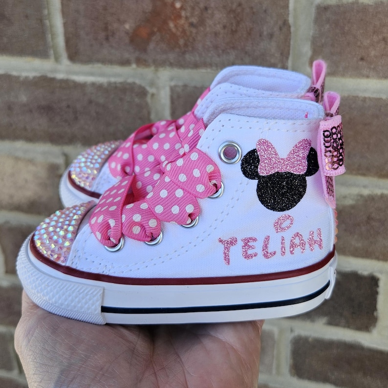 Puede incluir: Zapatillas blancas de ca&ntilde;a alta con cordones de cinta de lunares rosas y un dise&ntilde;o de Minnie Mouse con purpurina rosa. Las zapatillas est&aacute;n personalizadas con el nombre "Teliah".