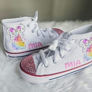 May include: White high-top trainers with pink rhinestone-covered toes. The trainers feature a colourful castle and Mickey Mouse design, with the name "MIA" in pink. The shoes have white laces and a red and blue stripe on the sole.