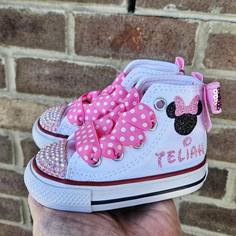 Puede incluir: Zapatillas blancas de ca&ntilde;a alta con cordones de cinta de lunares rosas, un dise&ntilde;o de Minnie Mouse con purpurina rosa y el nombre "YELIAN" en purpurina rosa en el lateral.